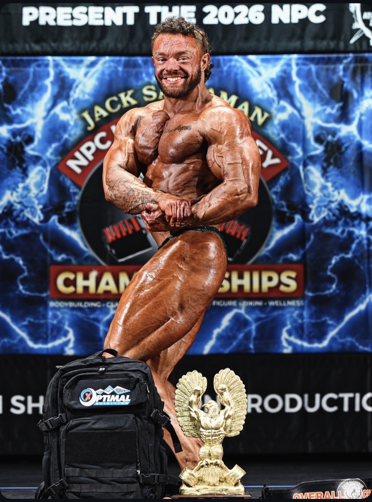 Jack Singleton, MVP coach, posing onstage with an overall champion trophy at an NPC bodybuilding show